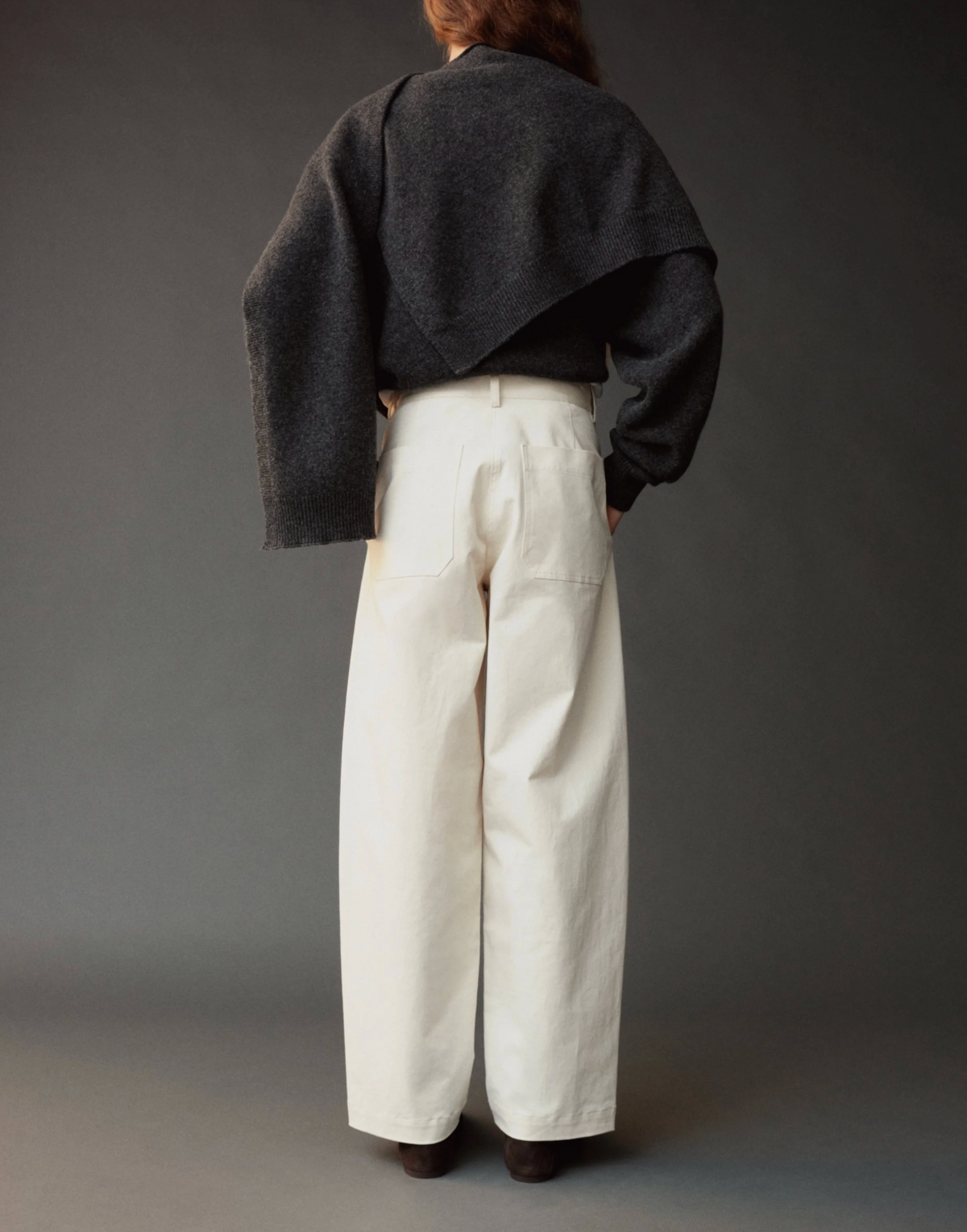 A model shown from the back wearing a dark charcoal cropped cardigan, a black long-sleeve top, and off-white wide-leg trousers against a dark gray studio background.