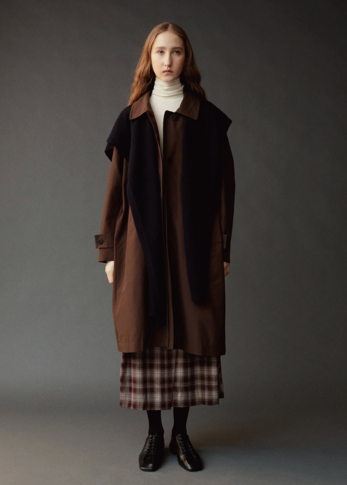 dark shawl over brown trench with plaid skirt and black shoes and white turtle neck on model