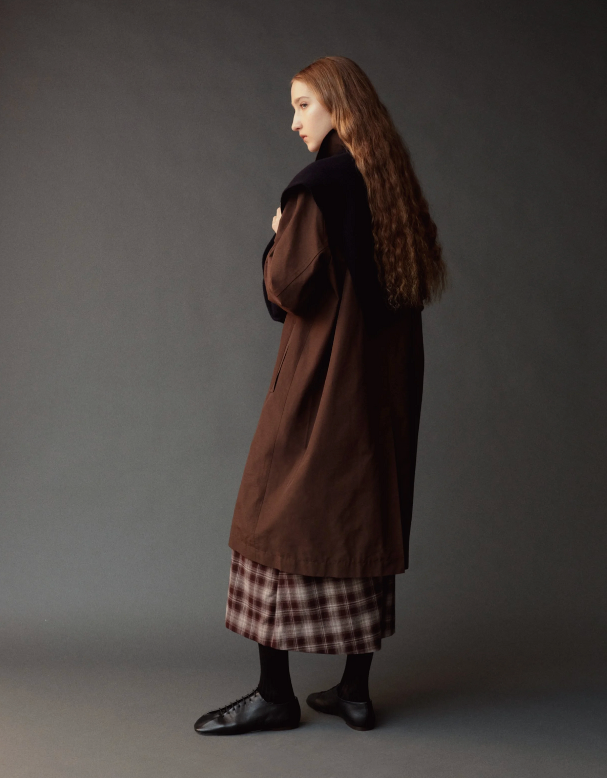 A model with long, wavy light brown hair is shown from the side profile, wearing a long brown coat over a brown and white plaid skirt, black tights, and black shoes, set against a dark gray studio background.