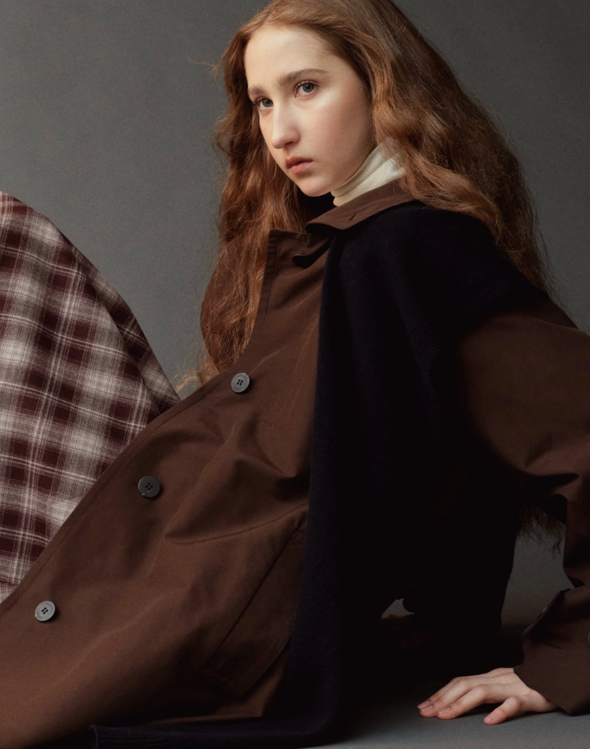 A model with long wavy light brown hair is wearing a dark brown double-breasted coat over a white turtleneck, with a dark navy woolen cape draped over her shoulders. She is posed against a dark gray studio background, with a plaid garment partially visible on the left.
