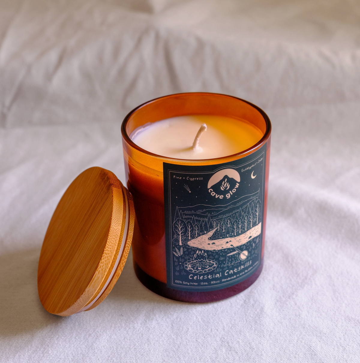 amber glass candle with wooden topper and green label with Cave Glow branding, info and drawing of the "celestial catskills," landscape with trees, fire, moon, mountains and more.