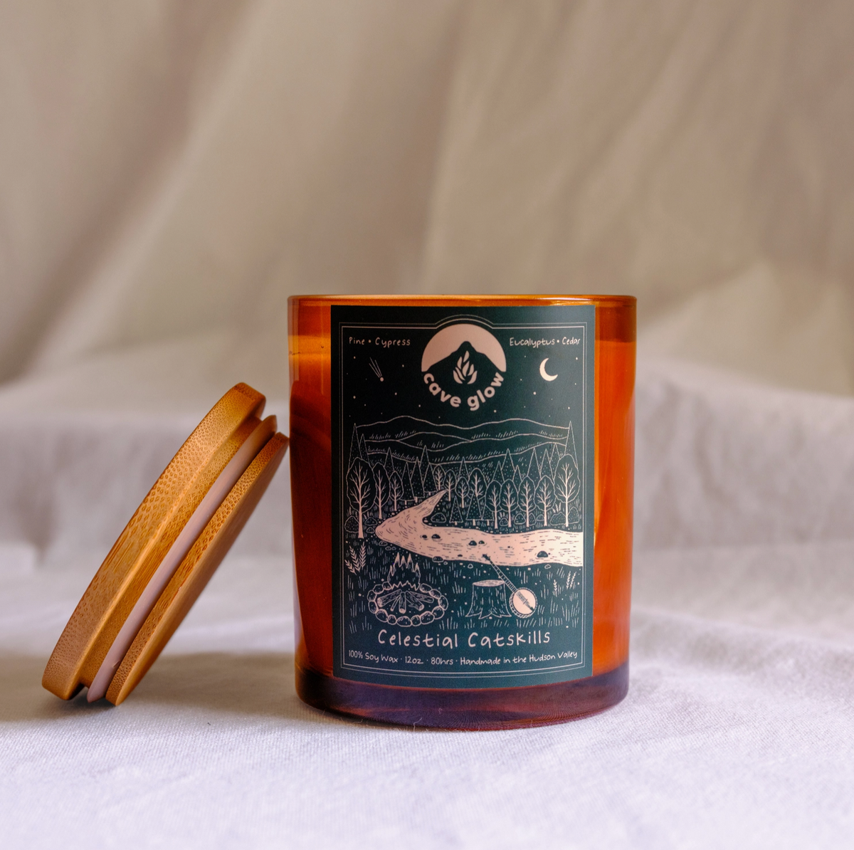 amber glass candle with wooden topper and green label with Cave Glow branding, info and drawing of the "celestial catskills," landscape with trees, fire, moon, mountains and more.