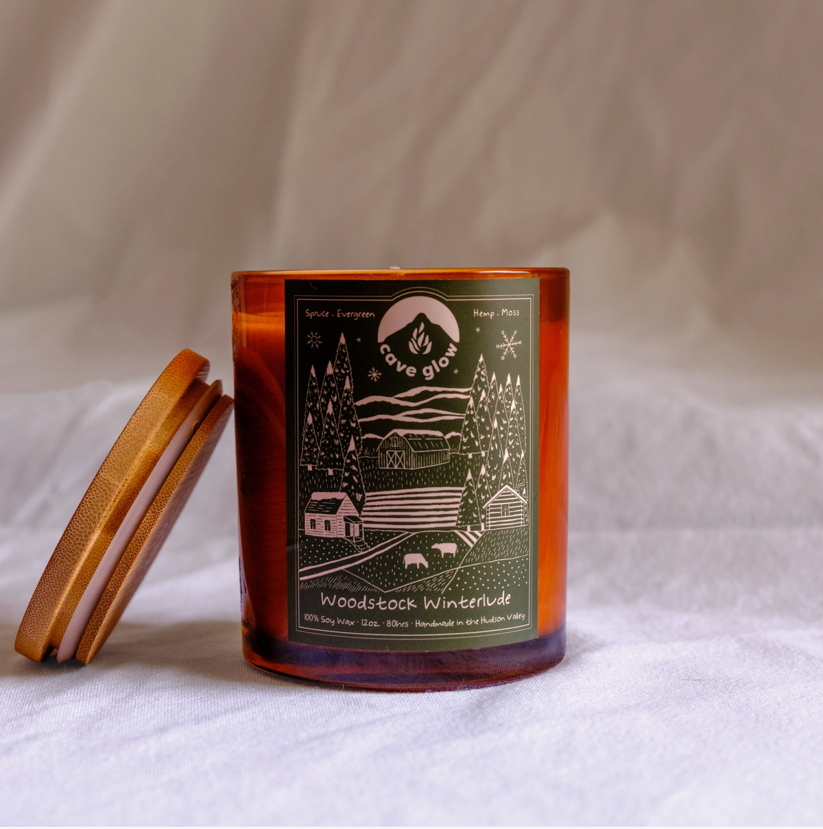 amber candle with wooden top, dark green label with branding and illustration of snowy outdoor landscape/farm with animals trees, a barn, a house, mountains and more