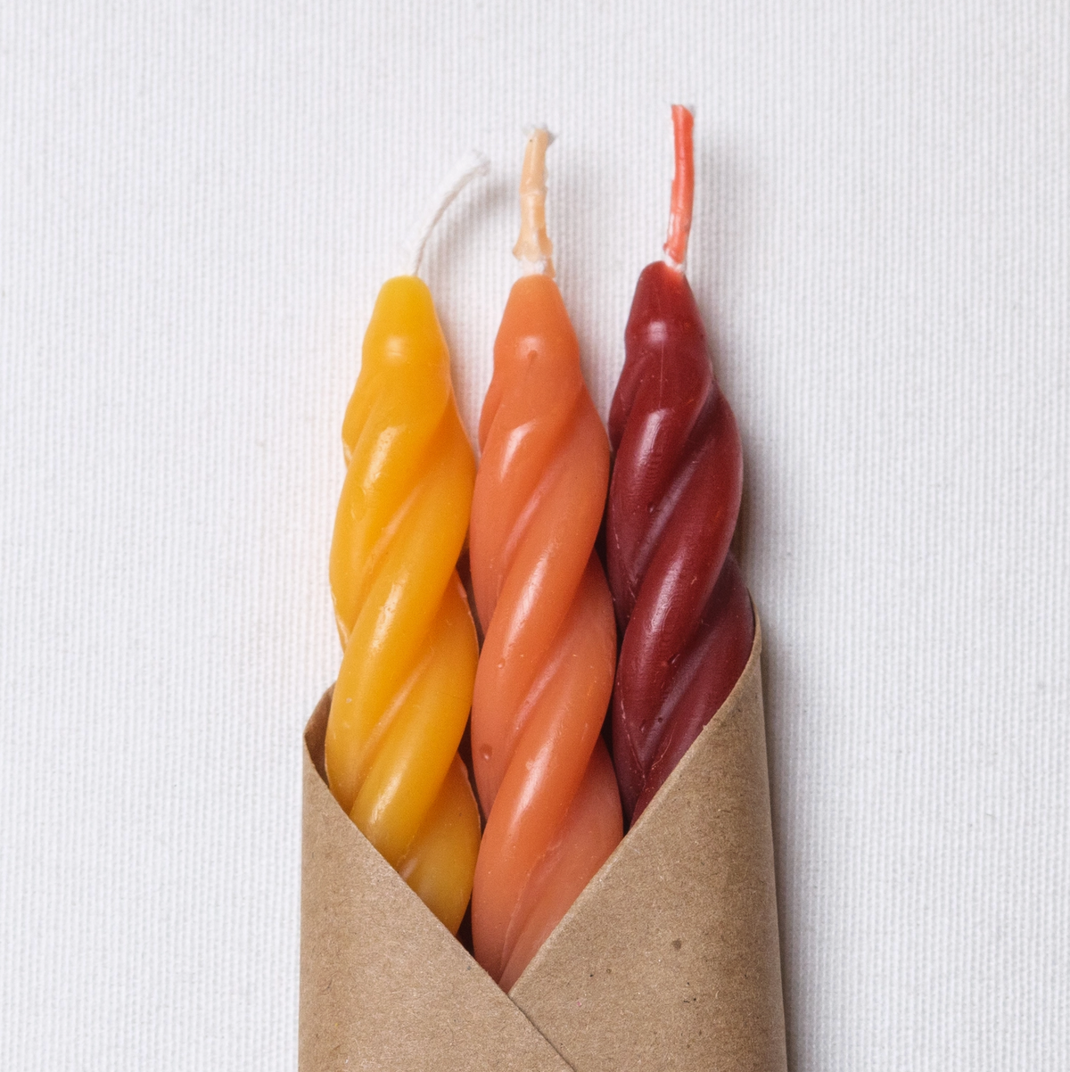 A package of three handcrafted wax candles in a brown kraft paper sleeve with a white label. The candles are twisted and come in three colors: yellow, orange, and red.