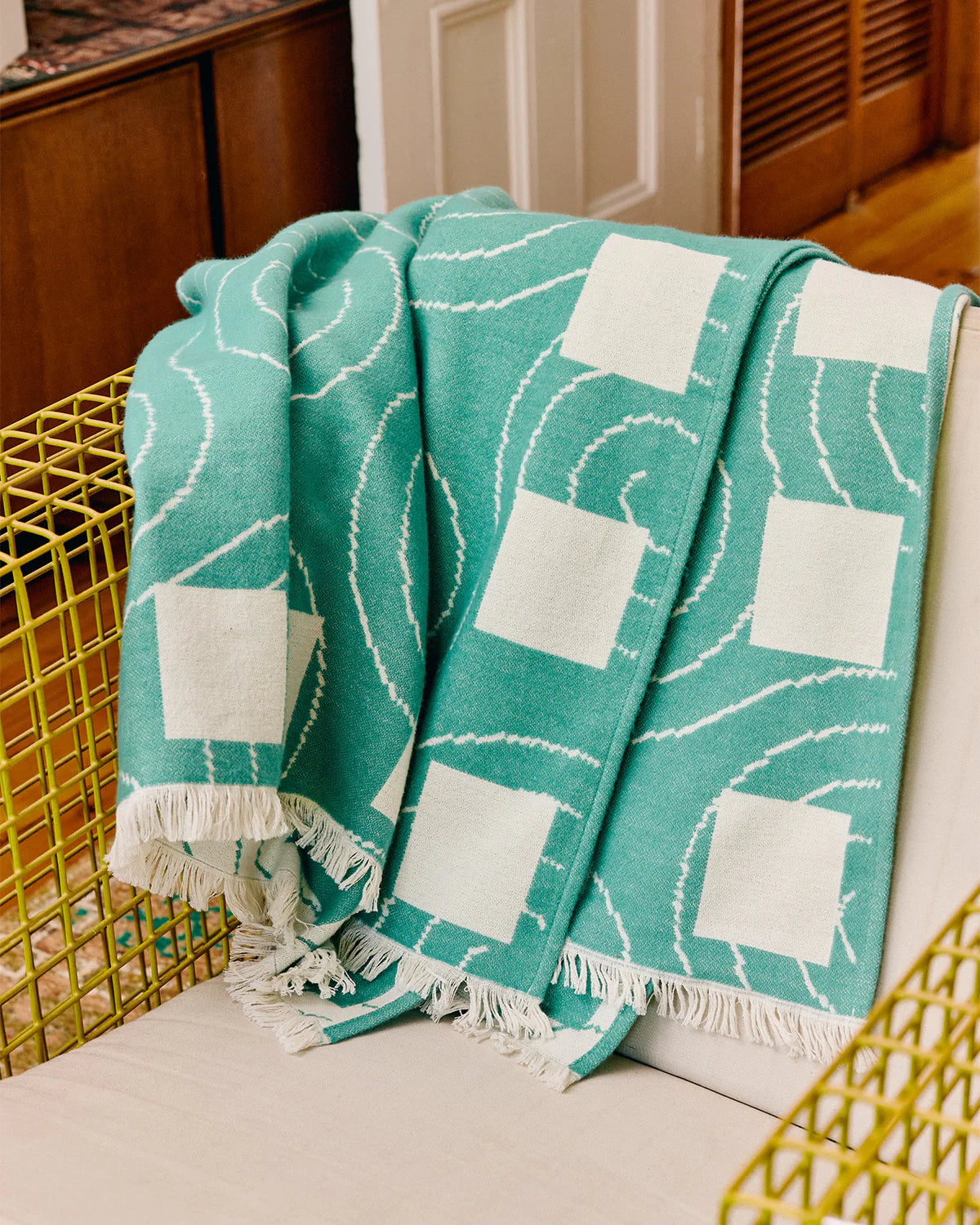 A teal and white patterned throw blanket with fringe is draped over a white chair in a home interior setting.