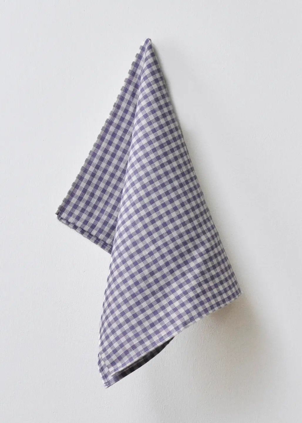 A small folded cloth napkin with a purple and white gingham check pattern, hanging against a plain white wall.