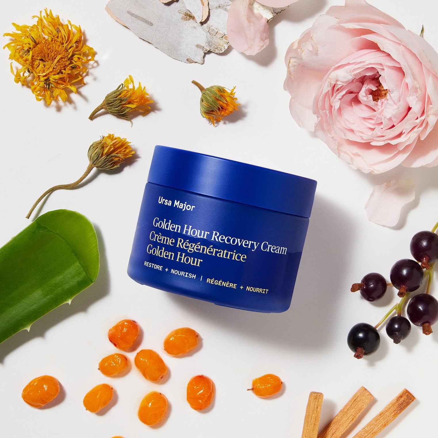 Blue jar of Ursa Major Golden Hour Recovery Cream surrounded by flowers and natural elements on a white background