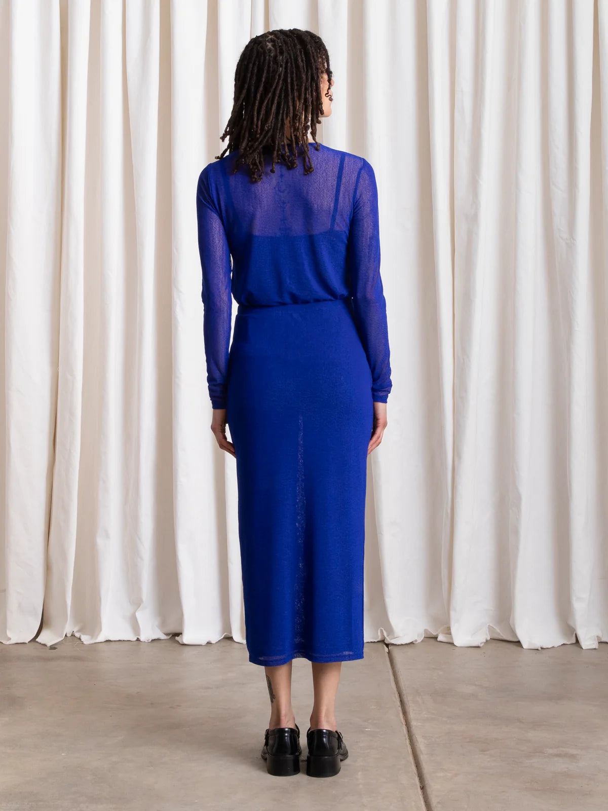 A model viewed from behind wearing a royal blue long-sleeve midi dress with a sheer mesh yoke, standing against a draped cream curtain backdrop.