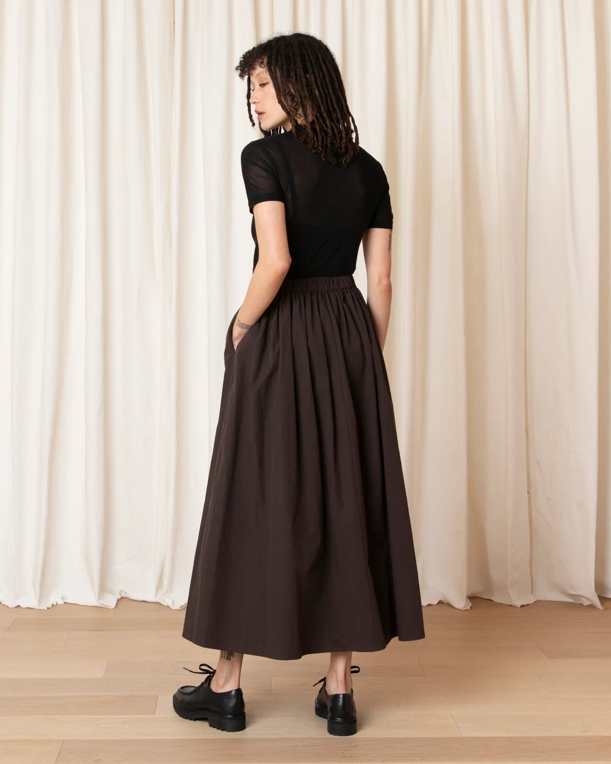 A woman stands with her back to the camera wearing a black short-sleeve mock-neck top and a dark brown pleated midi skirt with side pockets, paired with black platform shoes, set against a cream curtain backdrop.