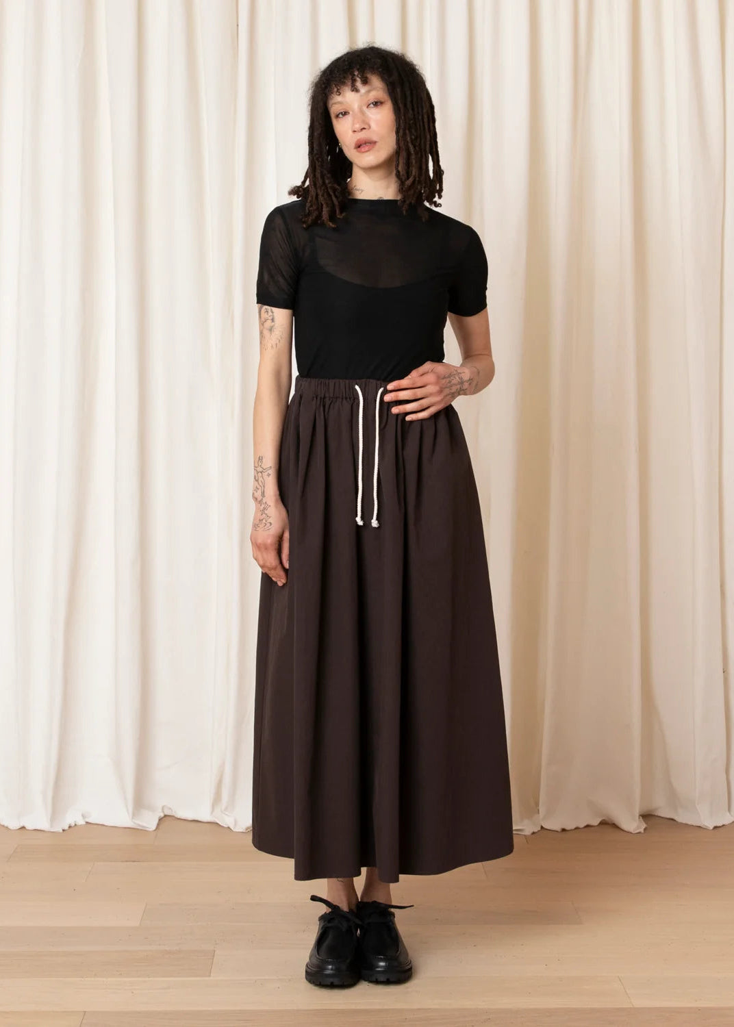 A model stands in front of cream-colored curtains wearing a black short-sleeve top with a sheer yoke and a long, wide-leg dark brown skirt with a white drawstring, paired with black shoes.