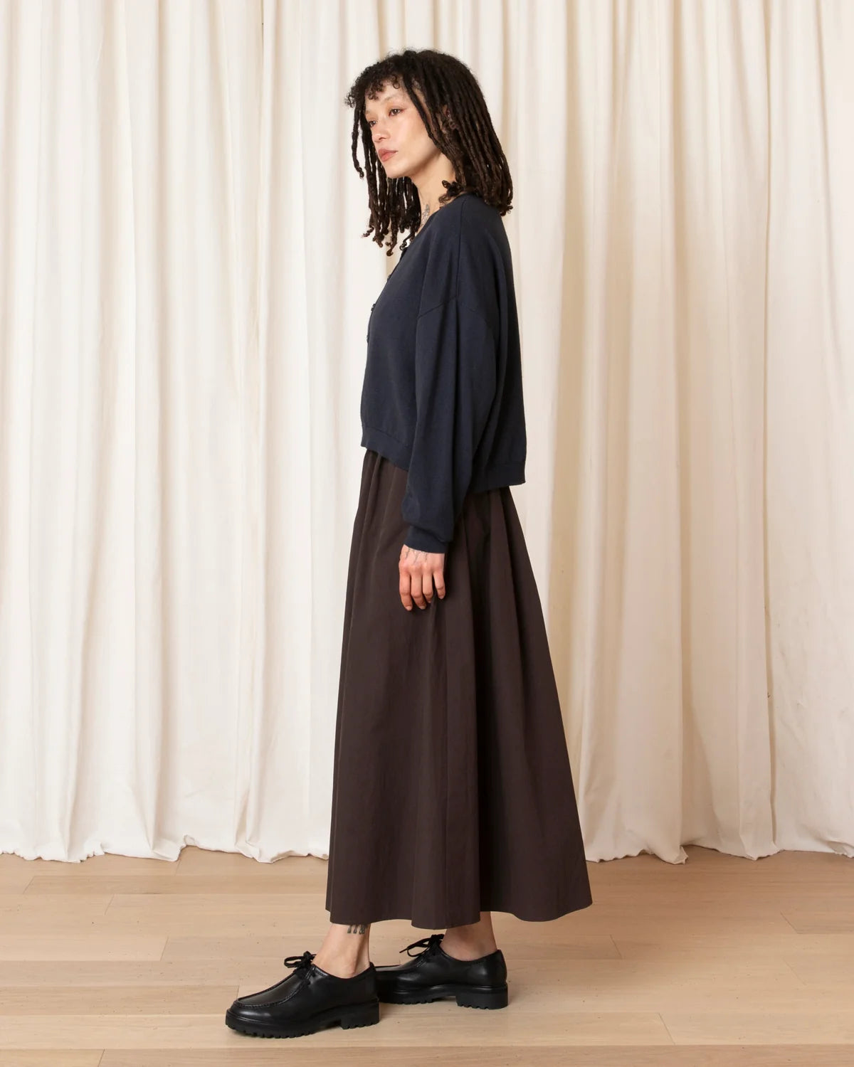 A woman stands in profile wearing a dark navy long-sleeve top and a dark brown midi skirt, paired with black leather shoes, set against a backdrop of draped cream curtains and a light wood floor.
