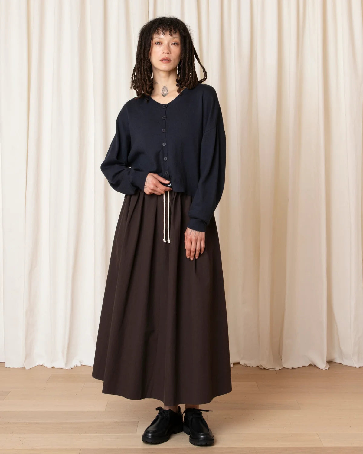 A model stands in front of a cream-colored curtain backdrop wearing a dark navy blue long-sleeve button-up top and a dark brown maxi skirt, paired with black leather shoes.