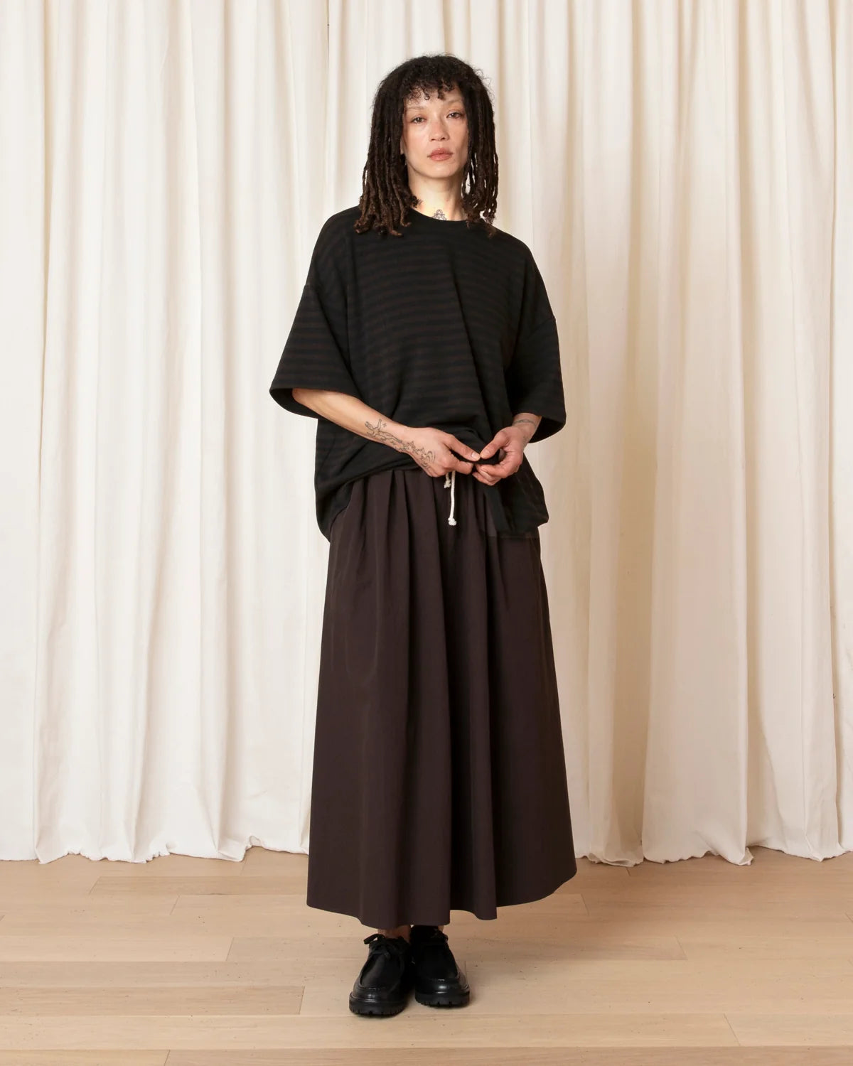 A model stands in front of a cream-colored curtain backdrop wearing a black ribbed short-sleeve top and a dark brown wide-leg skirt, paired with black shoes.