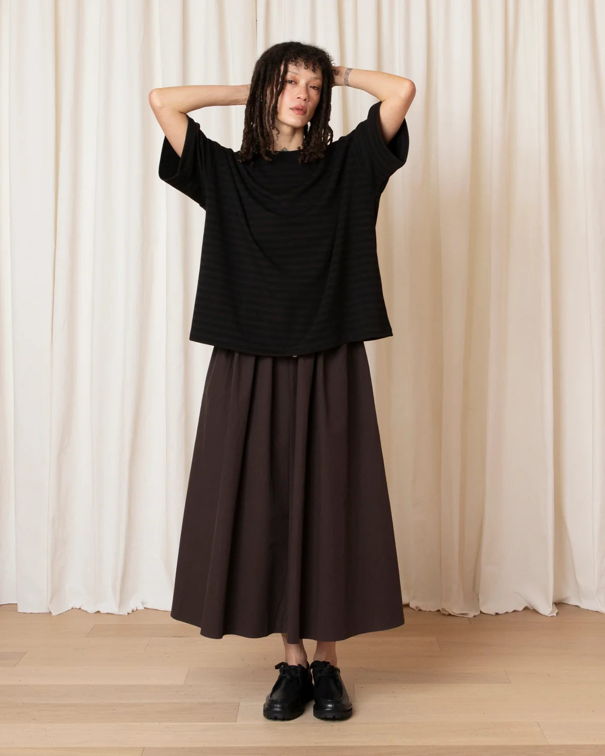 A model stands against a cream-colored curtain backdrop wearing a loose black short-sleeve top and a long dark brown pleated skirt, paired with black shoes.