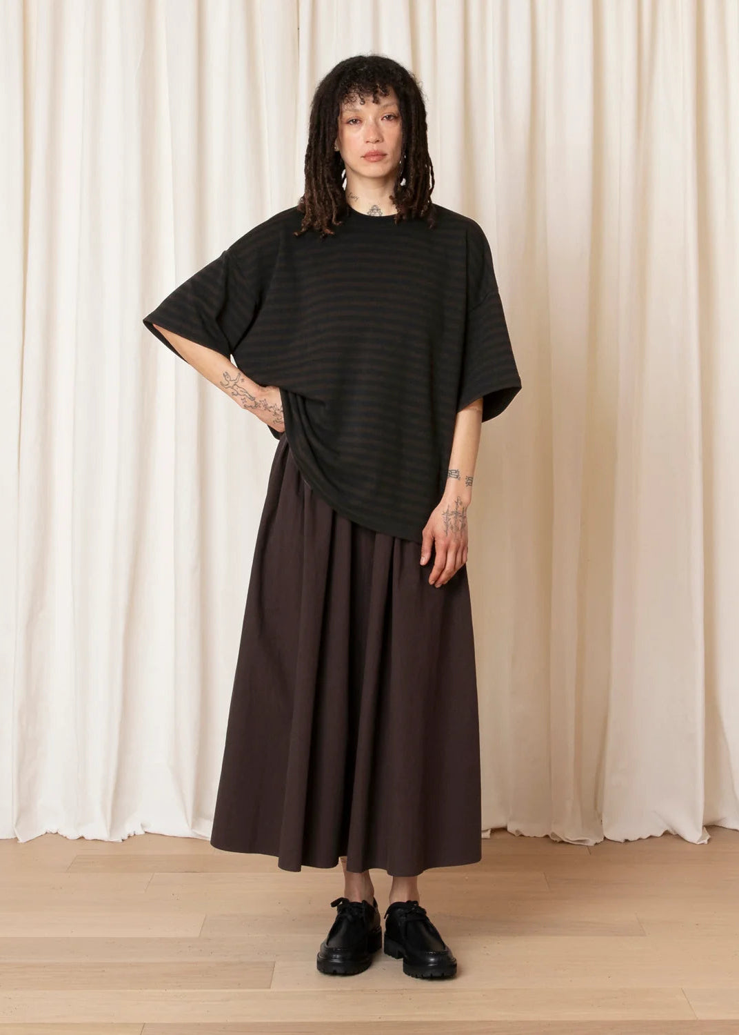 A model stands in front of a cream-colored curtain backdrop wearing a black and dark brown horizontally striped short-sleeve top, a dark brown pleated maxi skirt, and black leather shoes.