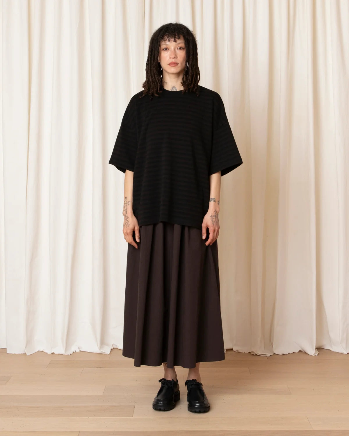A model stands in front of cream-colored draped curtains wearing a black short-sleeve striped knit top and a dark brown pleated maxi skirt, paired with black leather shoes.
