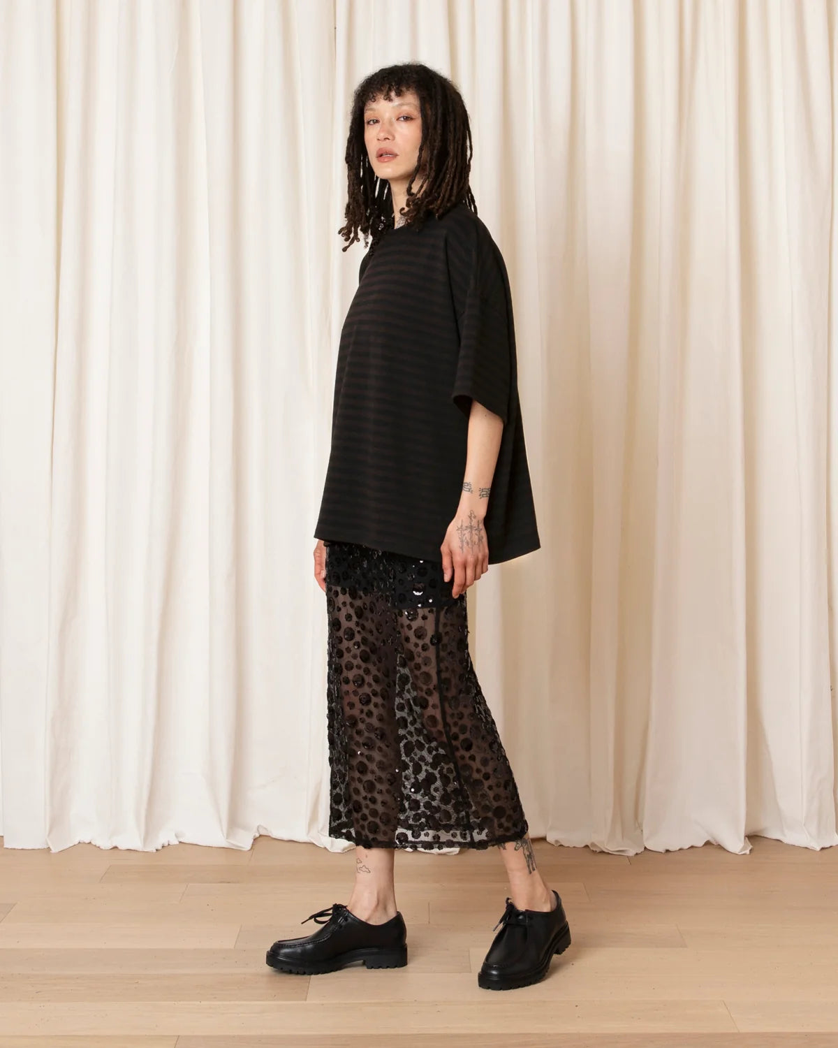 A model stands in profile wearing a loose black striped short-sleeve top, a sheer black leopard-print midi skirt, and black leather shoes, posed in front of a cream-colored draped curtain backdrop.
