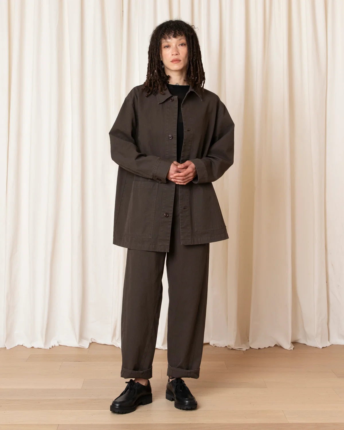 A model stands in front of a cream-colored curtain backdrop wearing a dark brown oversized button-up shirt jacket, matching dark brown trousers, a black top, and black leather shoes.