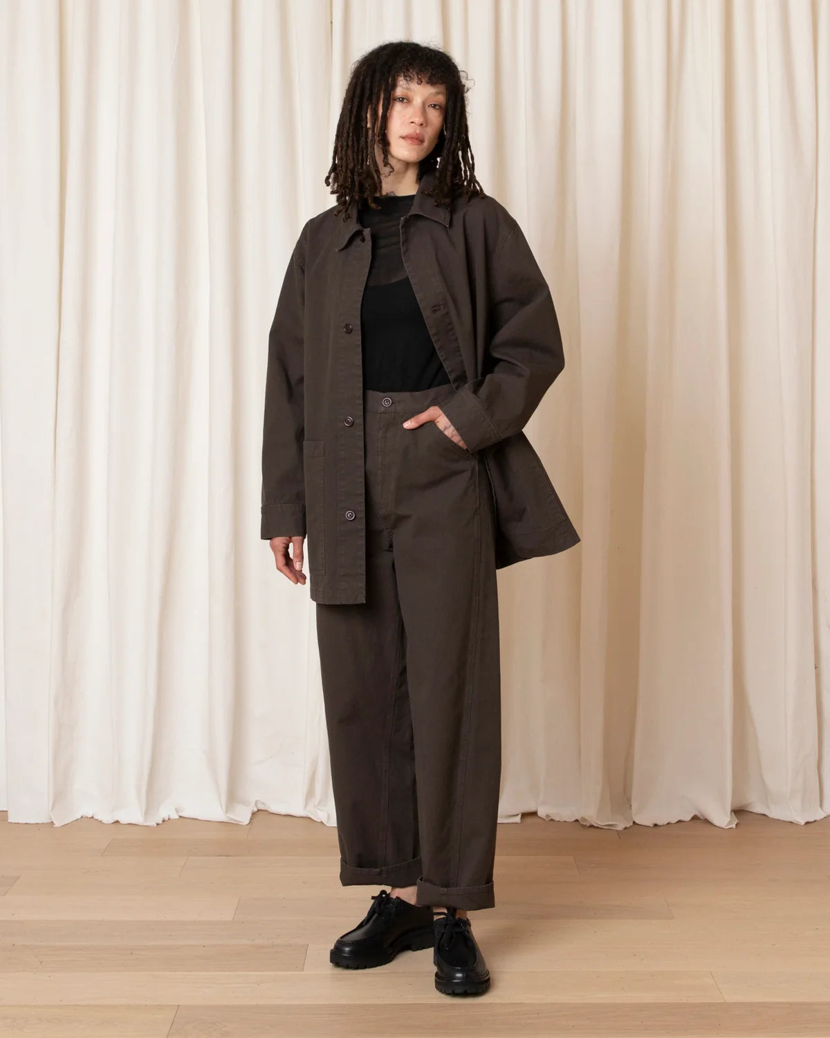 A model stands in front of cream-colored draped curtains wearing a dark brown oversized button-up jacket, matching wide-leg trousers, a black ribbed top, and black leather shoes.