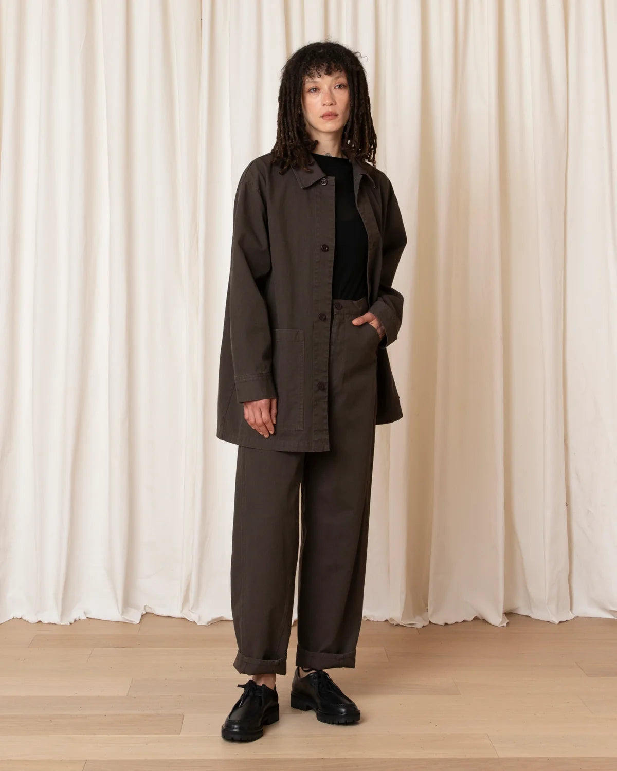 A model stands in front of a cream-colored curtain backdrop wearing a dark brown utility jacket, matching wide-leg trousers, a black top, and black leather shoes.