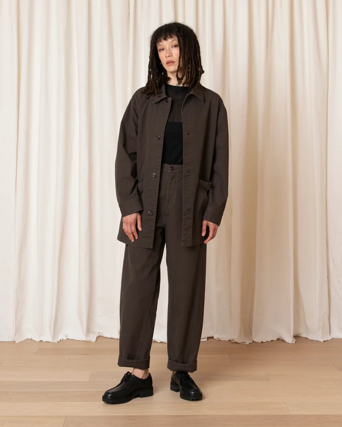 A model stands in front of a cream-colored curtain backdrop wearing a dark brown utility jacket, matching dark brown trousers, a black top, and black leather shoes.