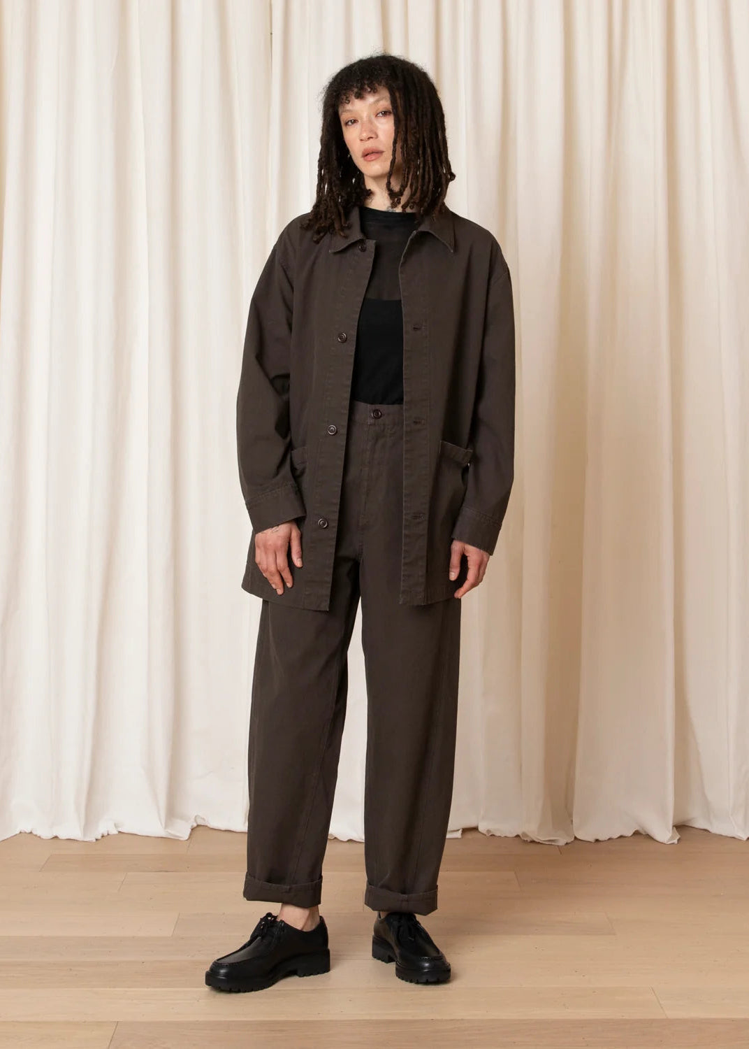 A model stands in front of a cream-colored curtain backdrop wearing a dark brown utility jacket, matching dark brown trousers, a black top, and black leather shoes.