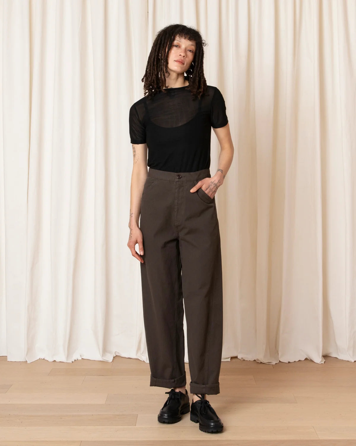 A model stands in front of a cream-colored curtain backdrop wearing a black short-sleeve mesh top and dark brown wide-leg trousers, paired with black shoes.