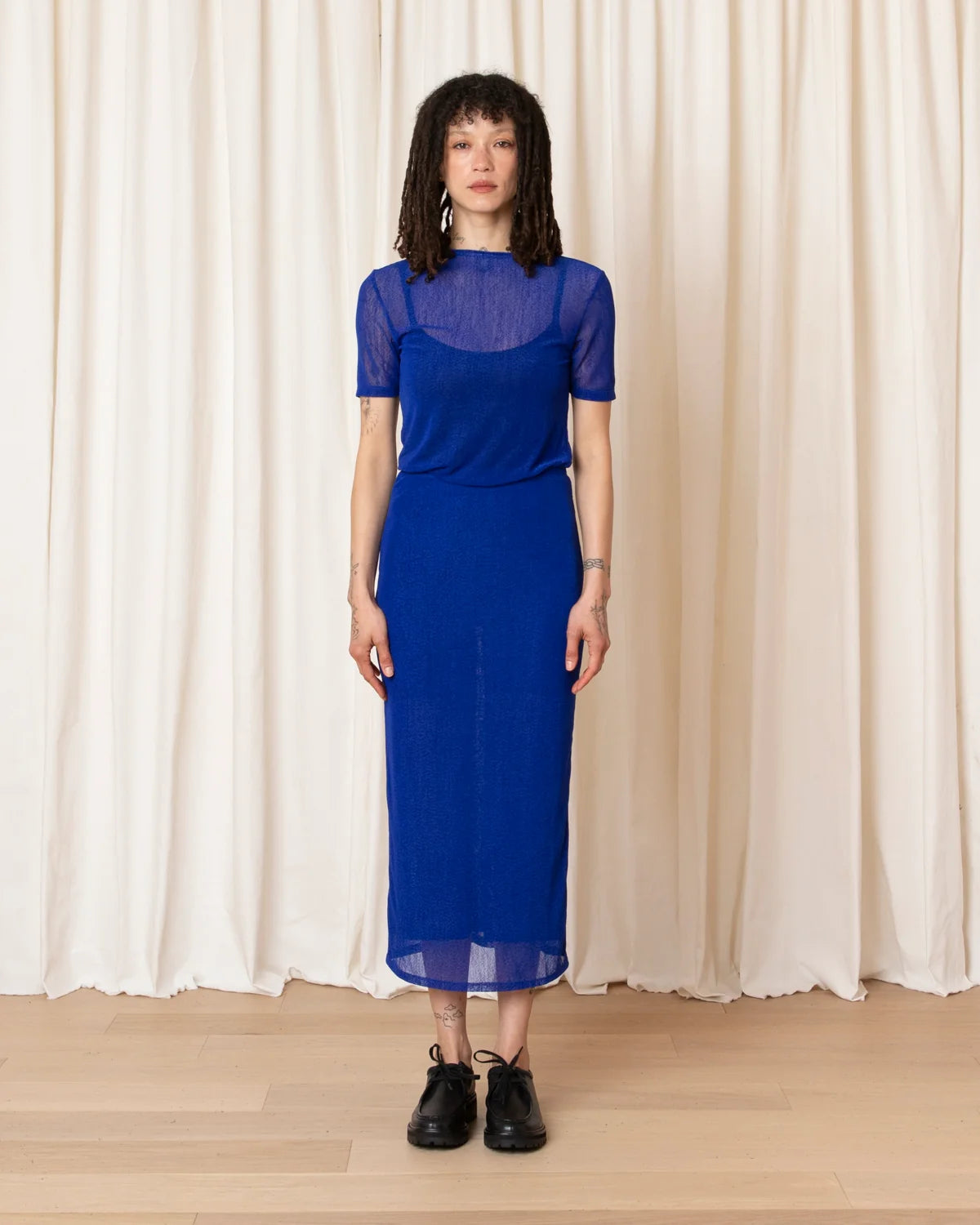 A woman stands in front of cream-colored draped curtains wearing a sheer royal blue midi dress with short sleeves and black lace-up shoes.