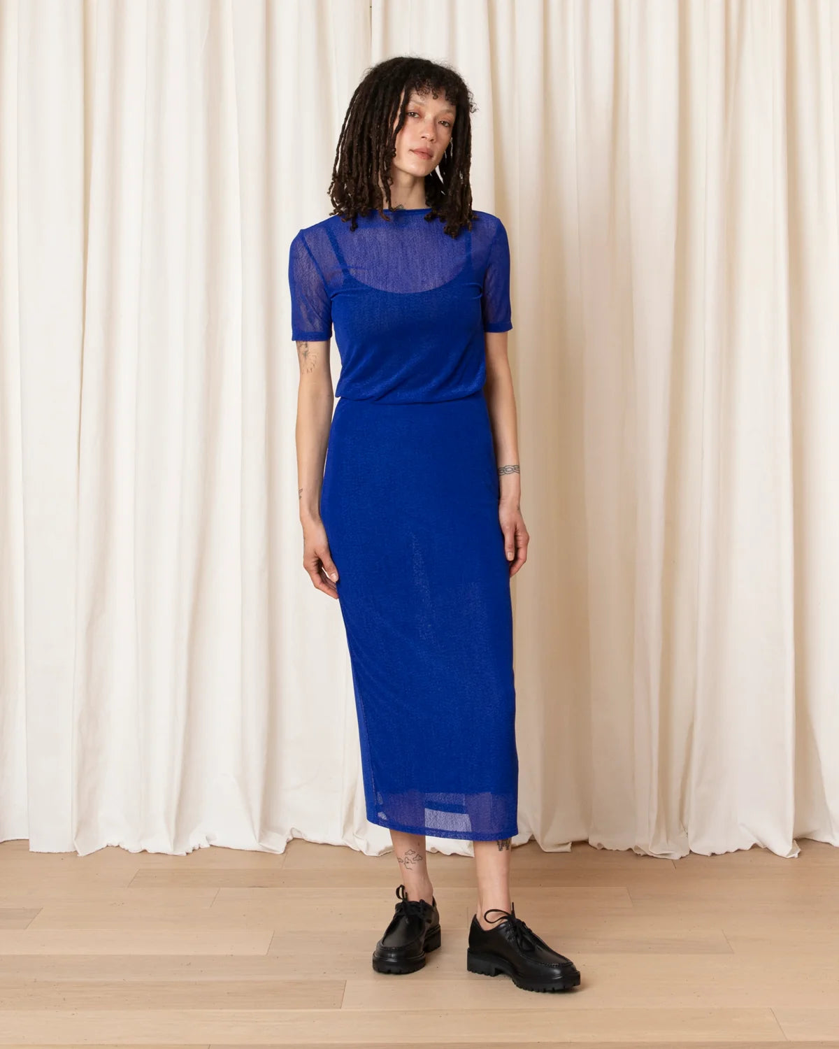 A woman stands in front of a cream-colored curtain backdrop wearing a vibrant royal blue midi dress with sheer short sleeves and a high neckline, paired with black leather shoes.