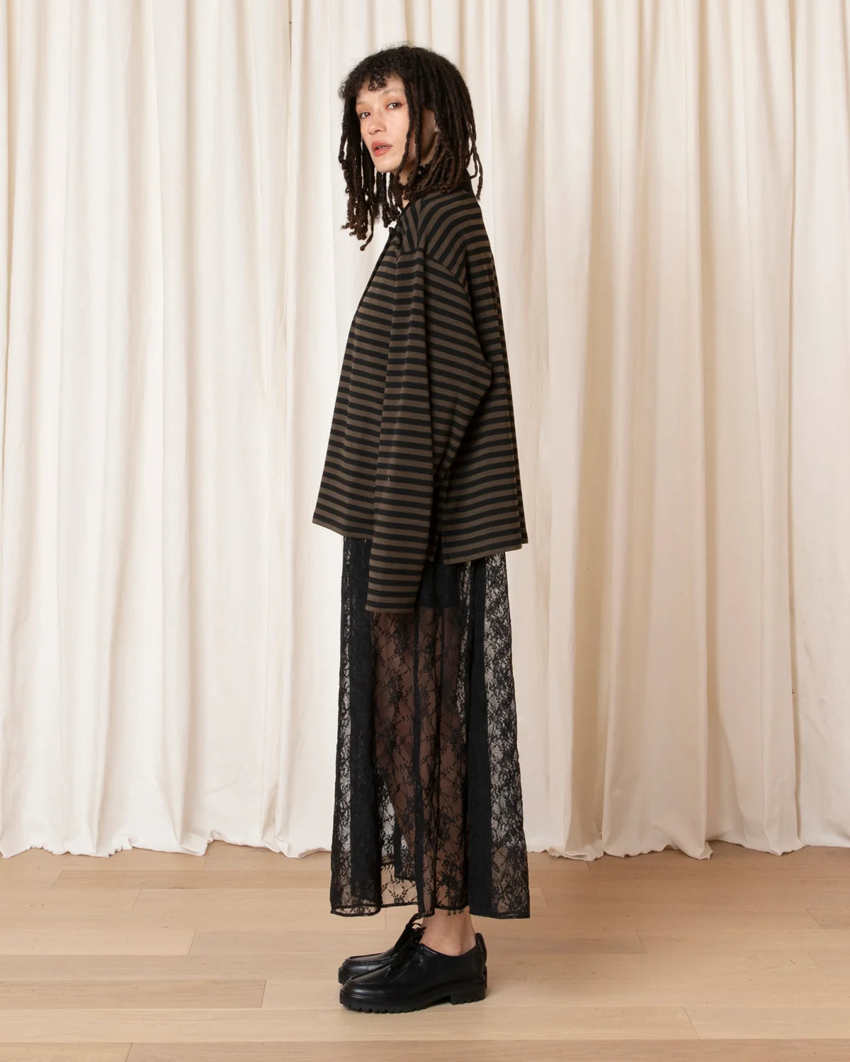 A model stands in profile wearing an oversized black and dark brown striped knit top over a long black lace skirt, paired with black leather shoes, set against a backdrop of cream-colored draped curtains.