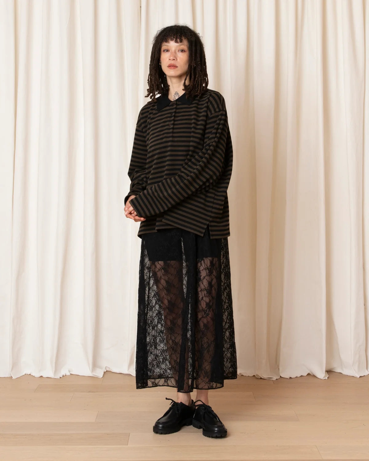 A model stands in front of cream-colored curtains wearing a black and dark brown striped long-sleeve top, a black lace overlay skirt, and black leather shoes.