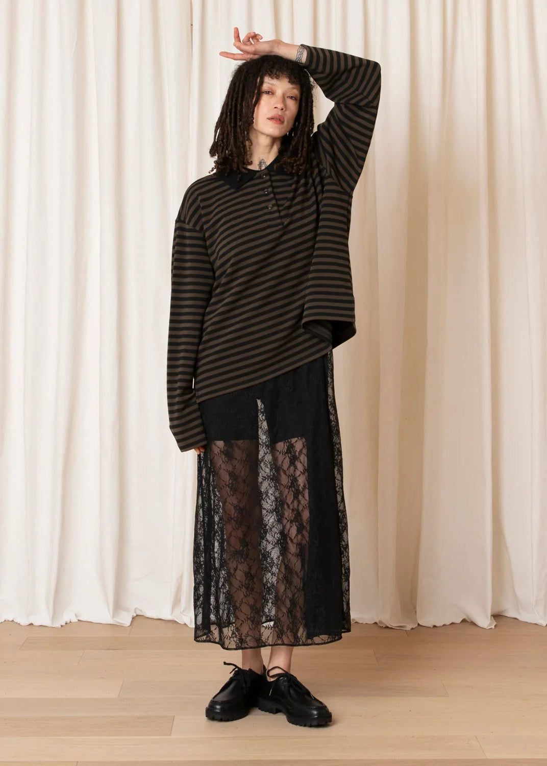 A model stands in front of cream-colored curtains wearing a black and dark brown striped long-sleeve top, a black midi skirt with a sheer floral lace overlay, and black leather shoes.
