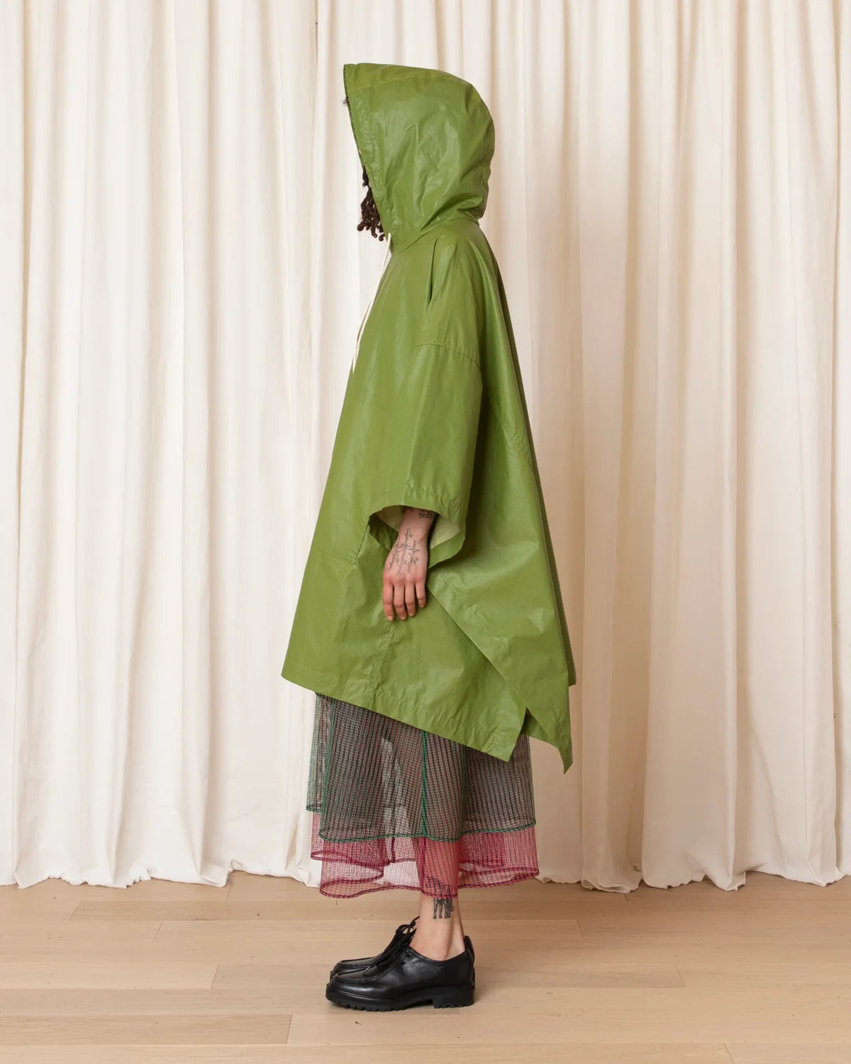 A person wearing an olive green hooded rain poncho over a striped skirt and black shoes, standing in front of cream-colored draped curtains.