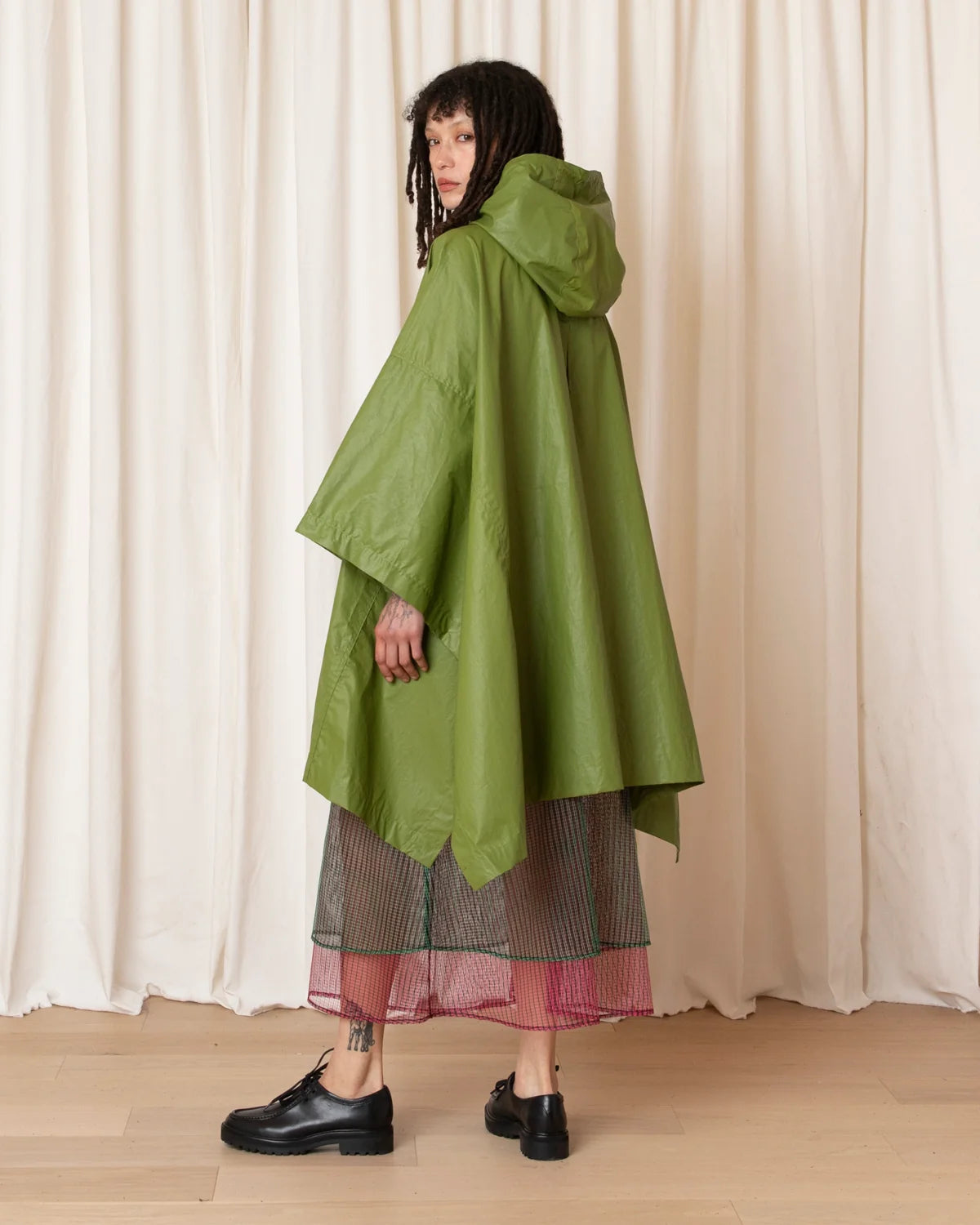 A model wearing green hooded rain poncho over a striped pink and gray skirt, standing in front of a cream-colored curtain backdrop.