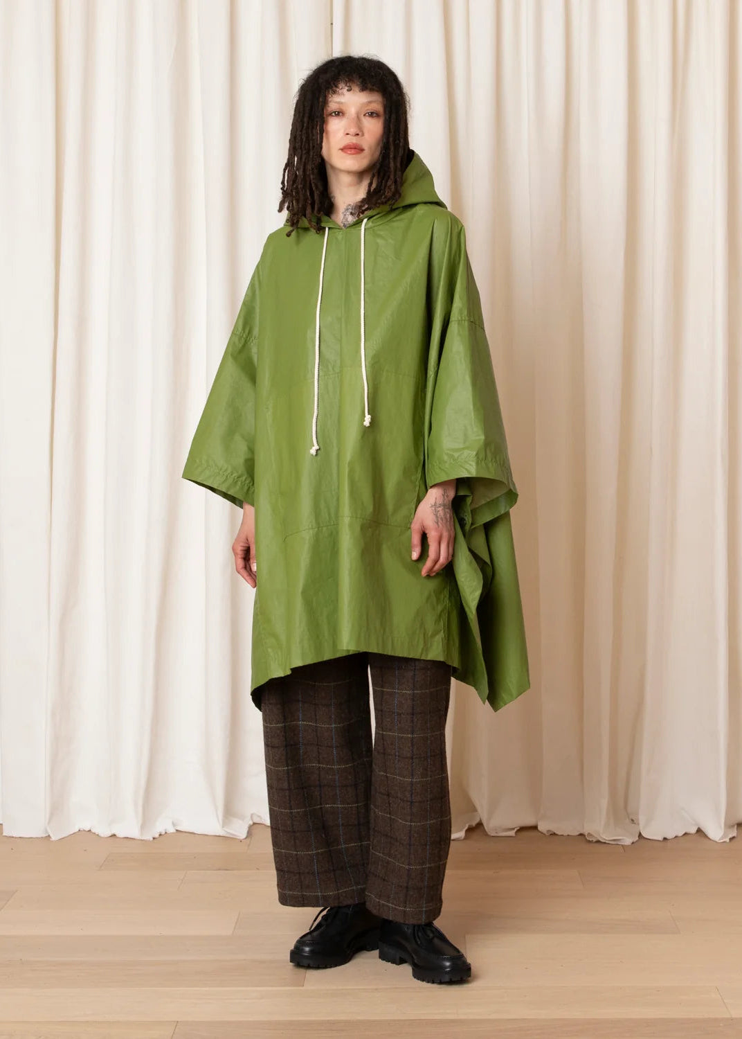 A model stands in front of cream-colored draped curtains wearing an olive green hooded rain poncho, brown plaid wide-leg trousers, and black shoes.