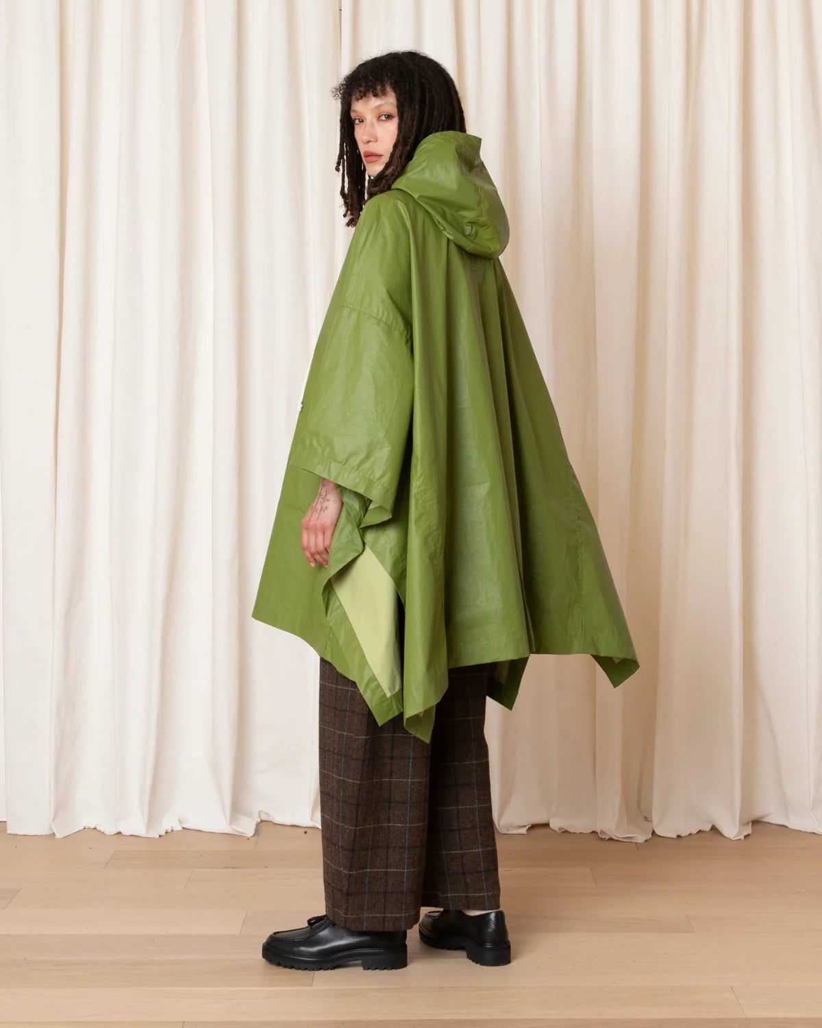 A model wearing green hooded rain poncho, brown plaid trousers, and black leather shoes, standing in front of a cream-colored curtain backdrop.