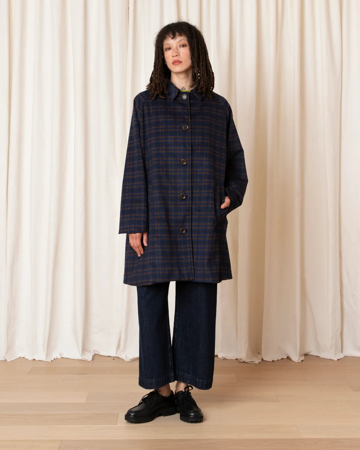 A model stands in front of a cream-colored curtain backdrop wearing a dark navy and red plaid coat, matching wide-leg trousers, and black leather shoes.