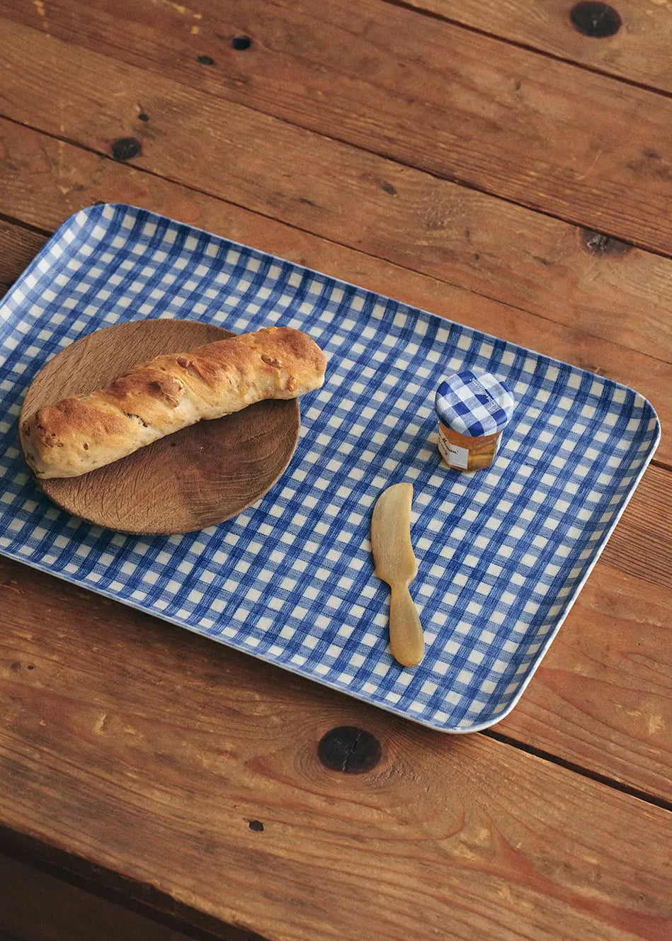 A rectangular blue-and-white gingham serving tray with rounded corners sits on a rustic wooden table. A loaf of bread rests on a small round wooden board on one side, and a small knife with a blue-and-white checkered handle lies on the tray.
