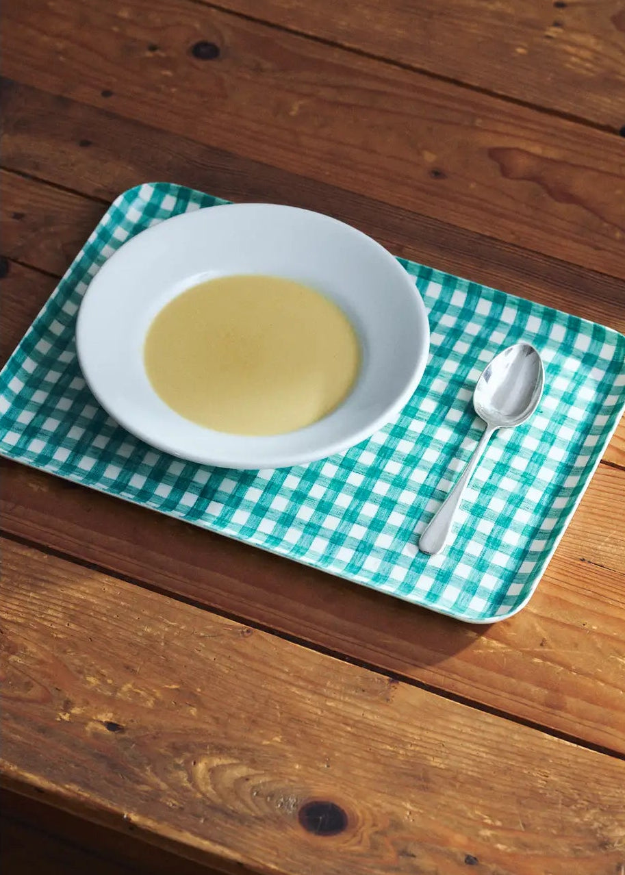 White bowl with yellow liquid on a green checkered tray with a spoon on a wooden table