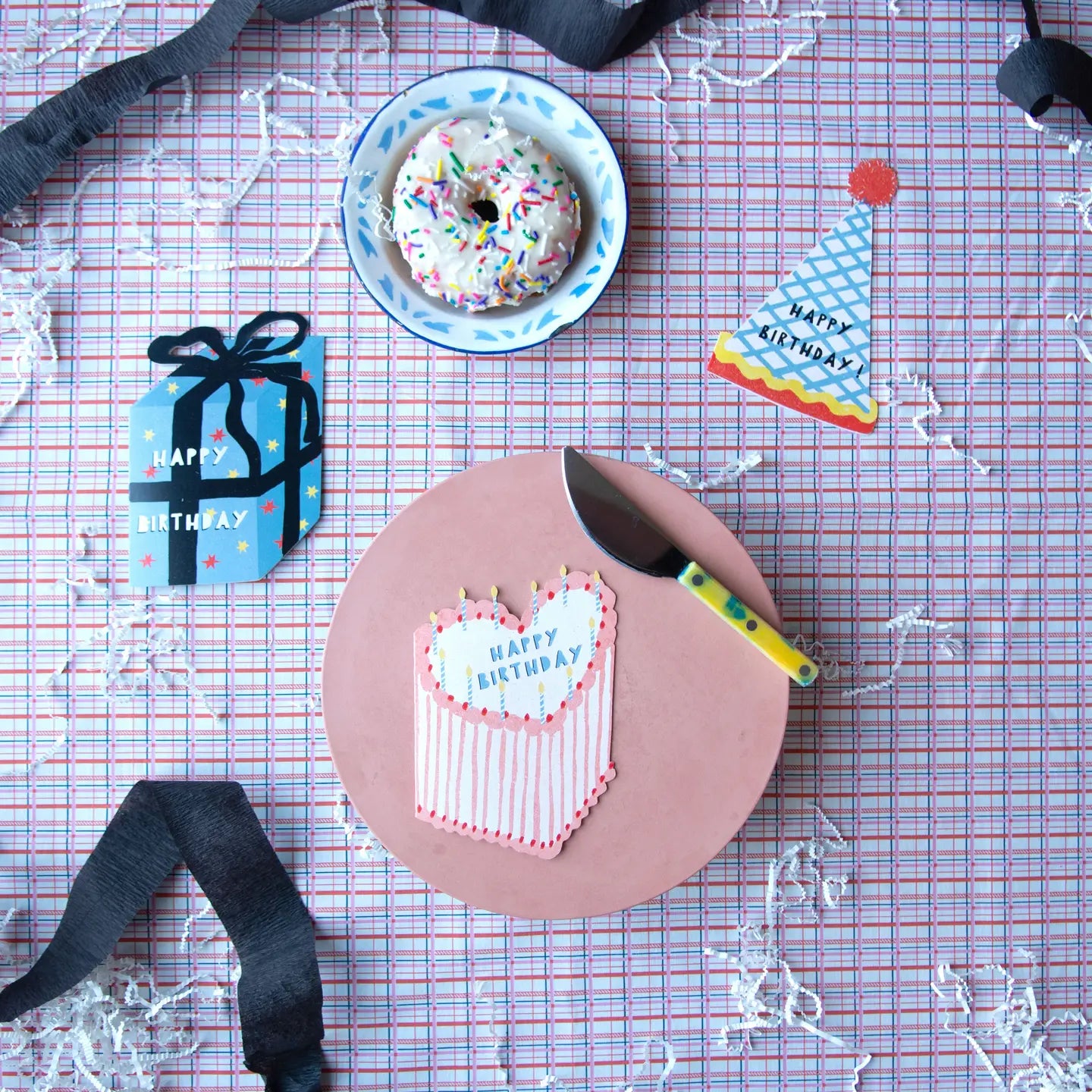 Birthday cake with 'Happy Birthday' decoration, knife, and donut on a checkered tablecloth.