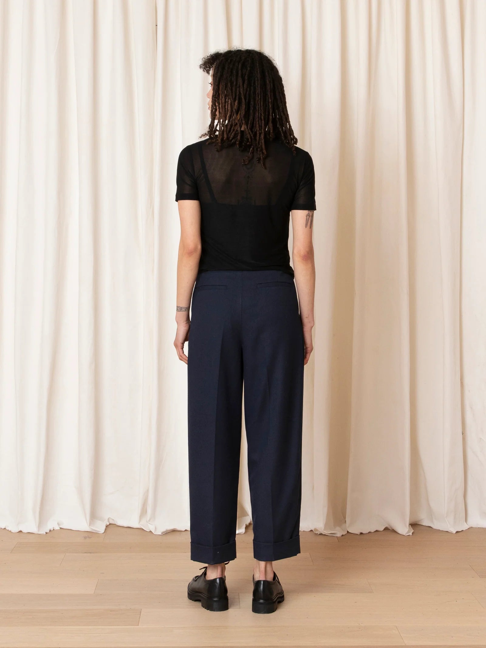 A model viewed from the back wearing a black sheer short-sleeve top and navy wide-leg cropped trousers, standing against a cream draped curtain backdrop.