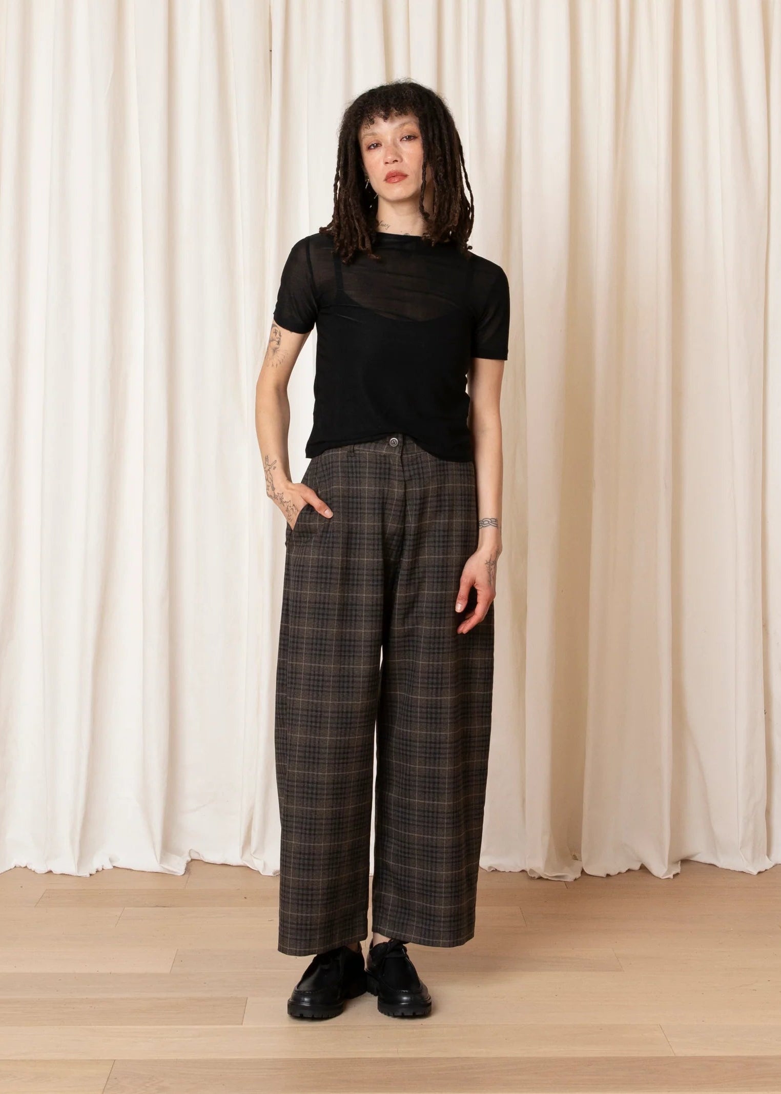 Person wearing a black top and plaid pants standing against a white curtain.