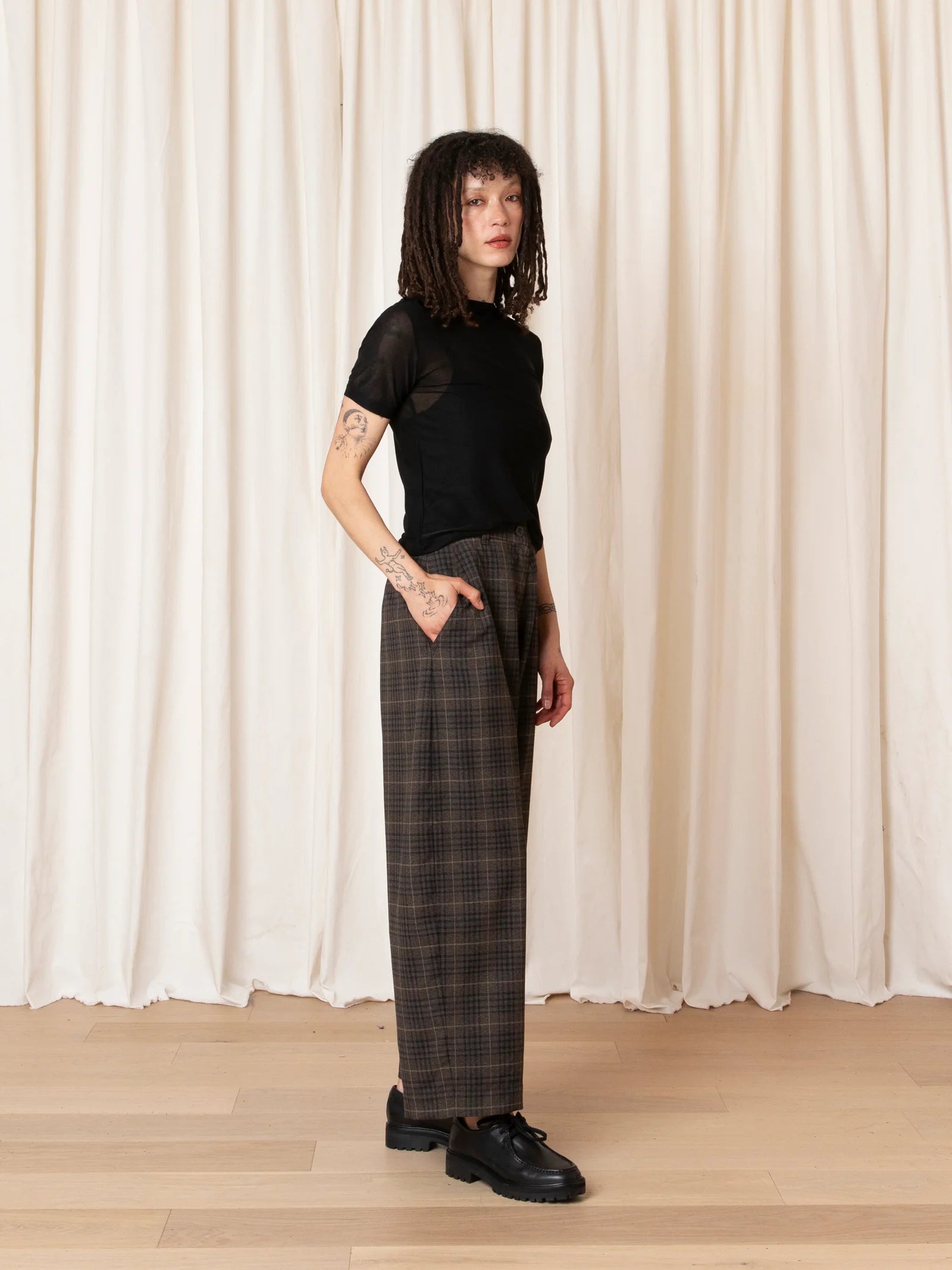 Person wearing a black top and plaid pants standing against a white curtain.