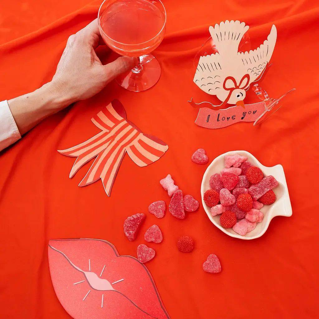 A styled flat-lay on a bright orange-red fabric background featuring Valentine's themed paper cutouts (lips, bow, dove with 'I love you' banner), a small white fish-shaped dish filled with pink and red heart-shaped gummy candies, and scattered heart gummies. A hand holding a pink drink enters from the left.
