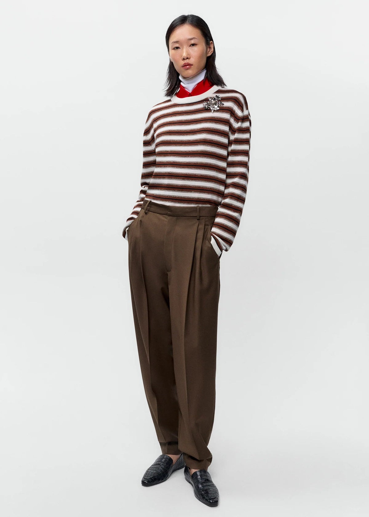 A model stands against a light gray studio background wearing a brown and white striped knit sweater with a red collar, brown pleated trousers, and black loafers.