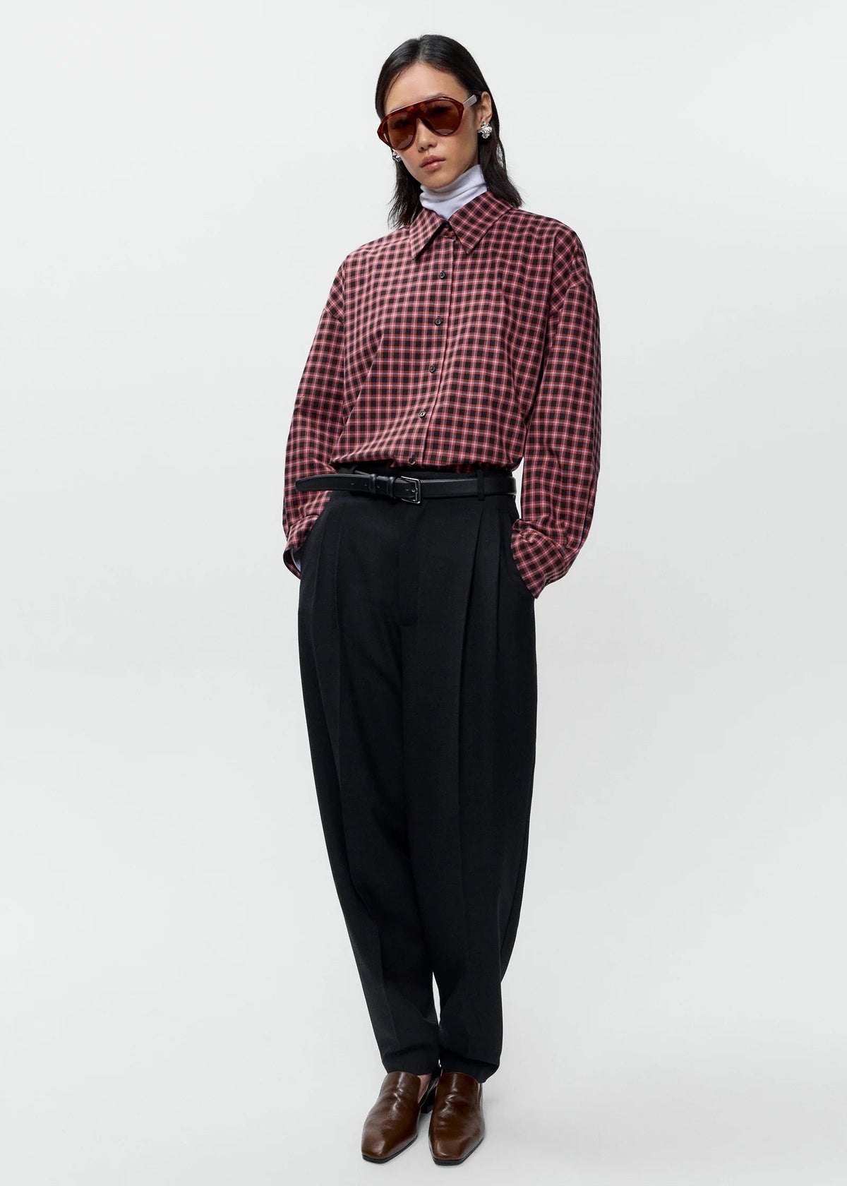 A model stands against a light gray studio background wearing a red and black checkered long-sleeve button-up shirt, black wide-leg trousers, a black belt, brown leather shoes, and dark sunglasses.
