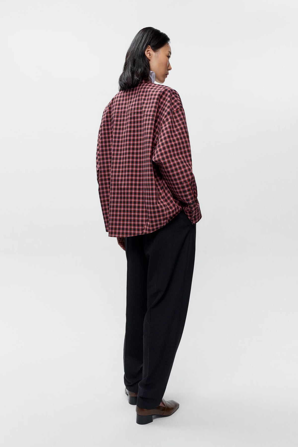 A model shown from the back wearing a red and black checkered plaid button-up shirt, black trousers, and brown leather shoes against a plain white studio background.