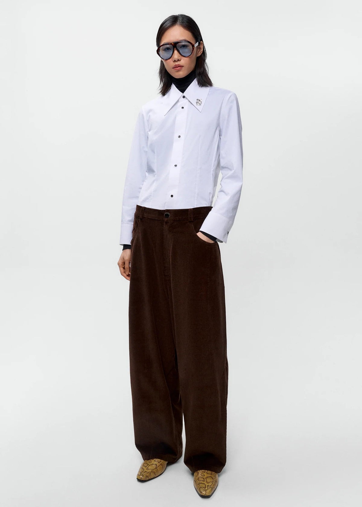 A model stands against a light gray studio background wearing a white long-sleeve button-up shirt, dark brown wide-leg corduroy trousers, patterned brown shoes, and dark sunglasses.