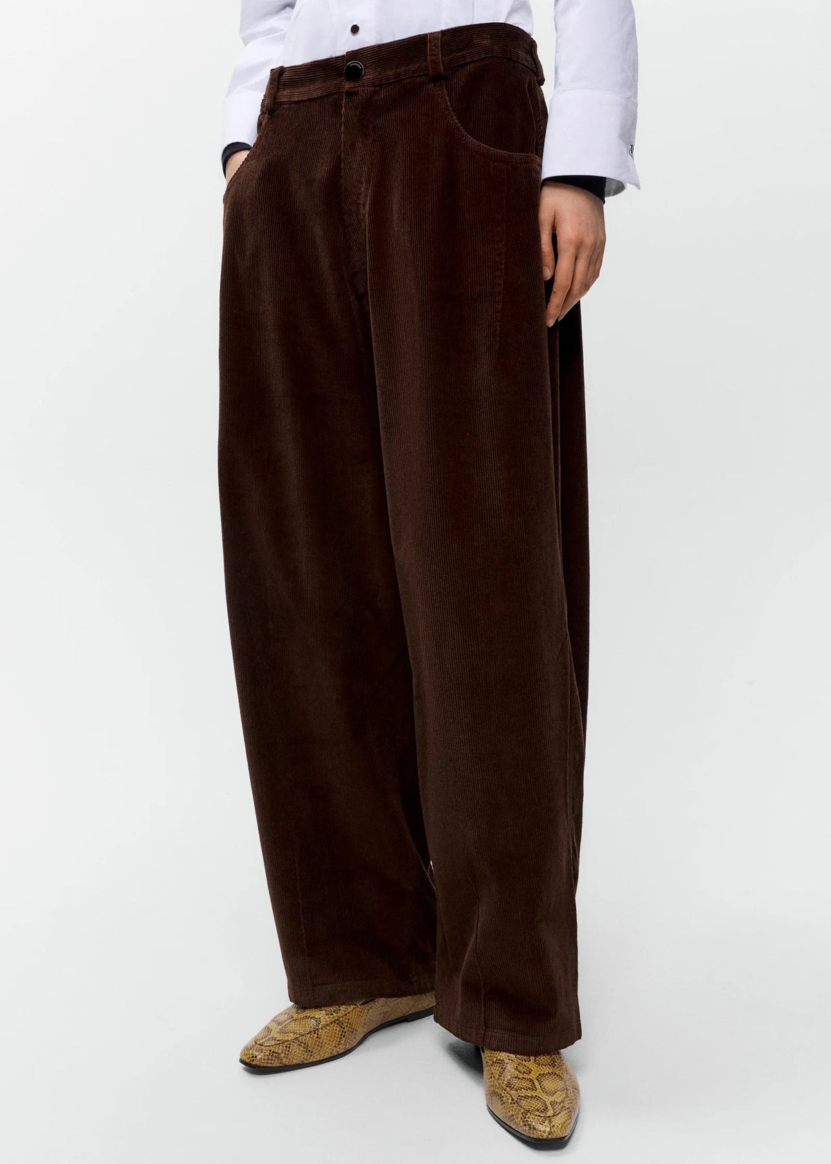 A person wearing very wide-leg dark brown corduroy trousers, styled with a white button-down shirt and brown patterned boots, photographed against a clean white studio background.