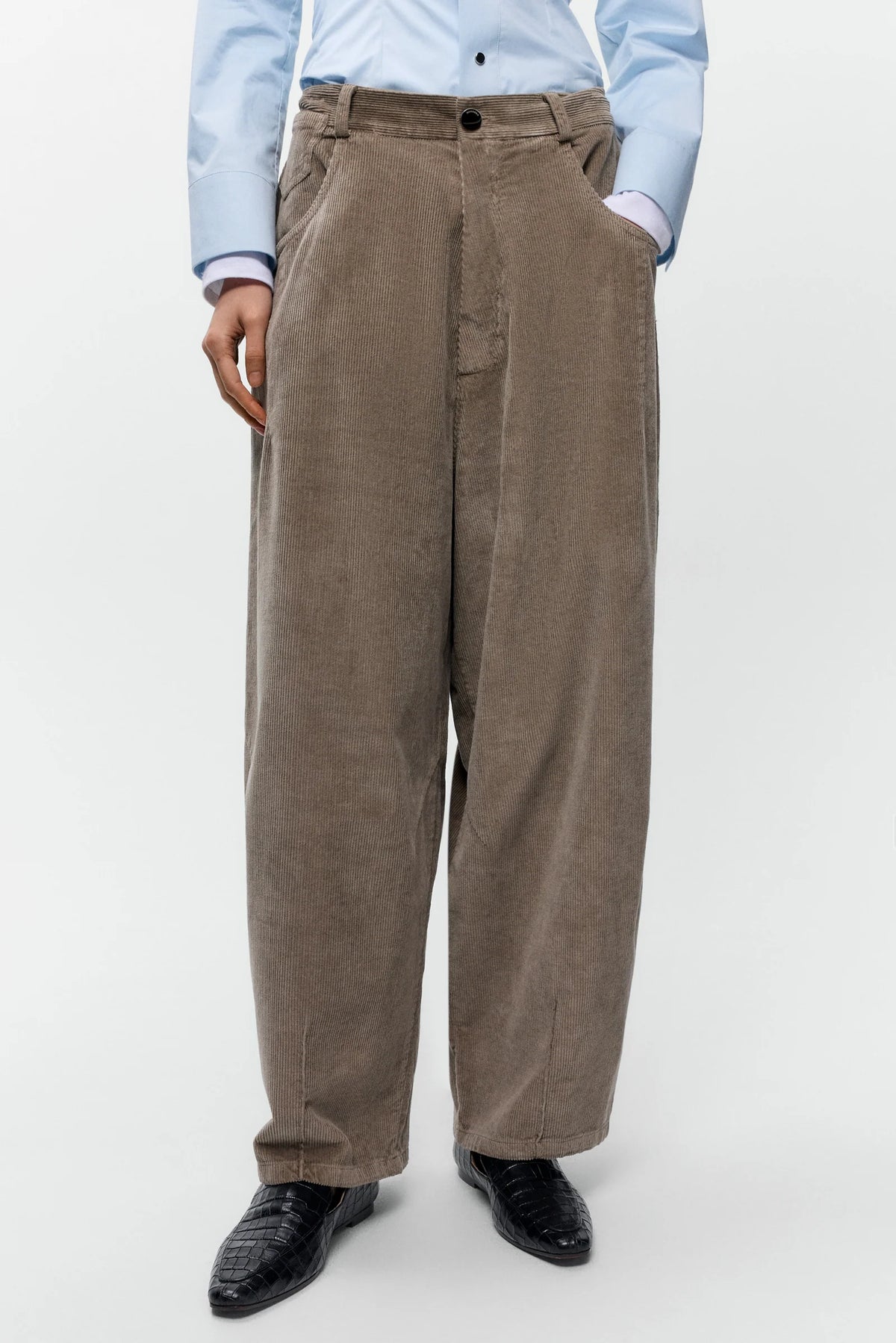 A person wearing very wide-leg taupe corduroy trousers, styled with a light blue dress shirt and black leather shoes, photographed against a clean white studio background.